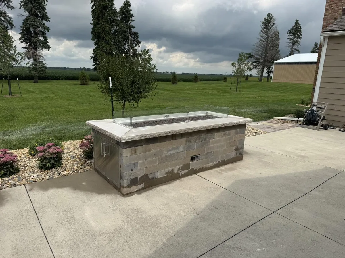 Outdoor fire table with glass wind guard, masonry base, and landscaped planting bed in Southern Minnesota